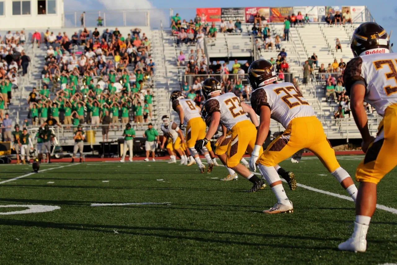 High school football game in action with stands of fans cheering in team colors under bright lights
