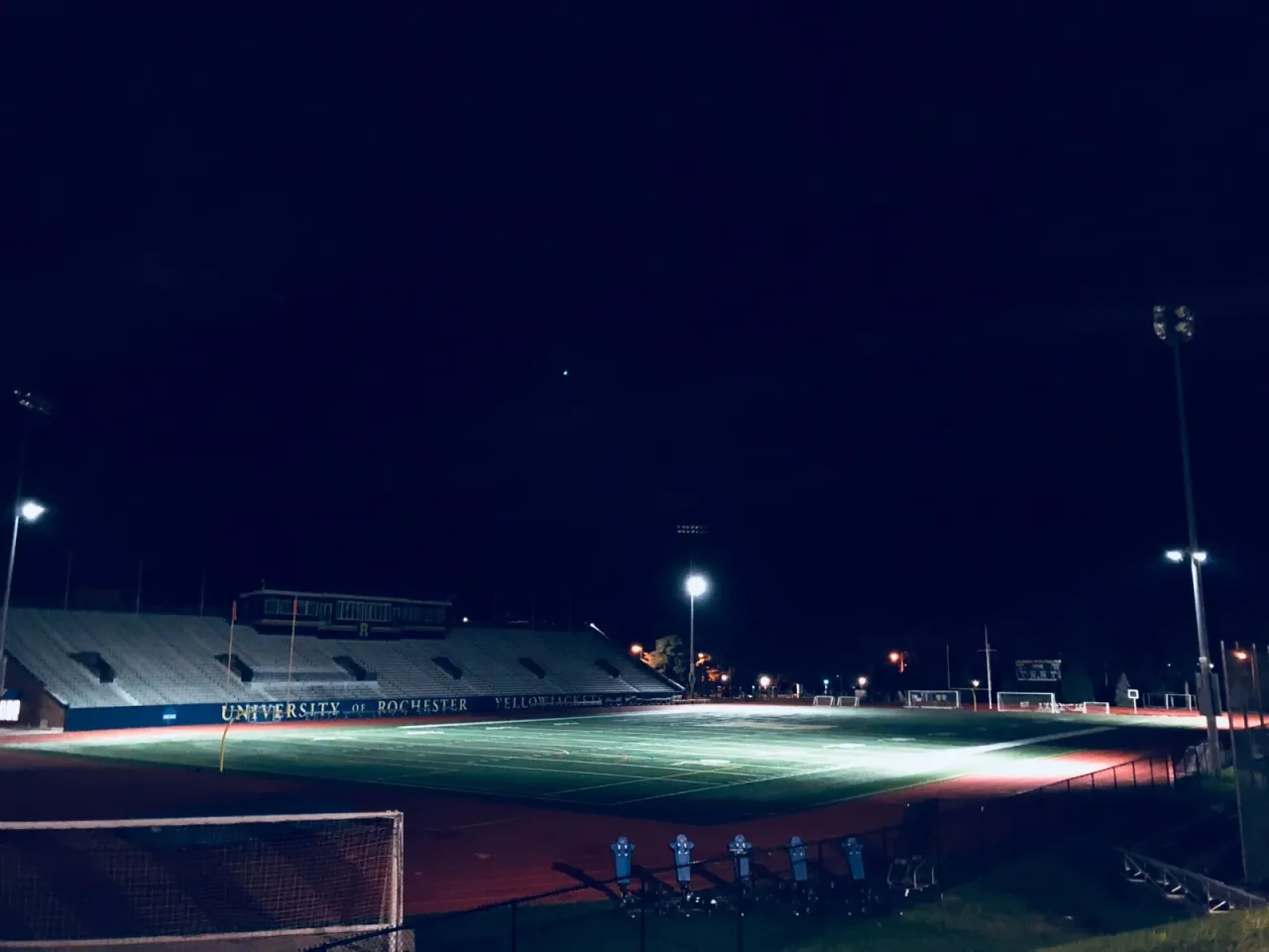 A small college stadium lit up at night with an empty field and surrounding bleachers