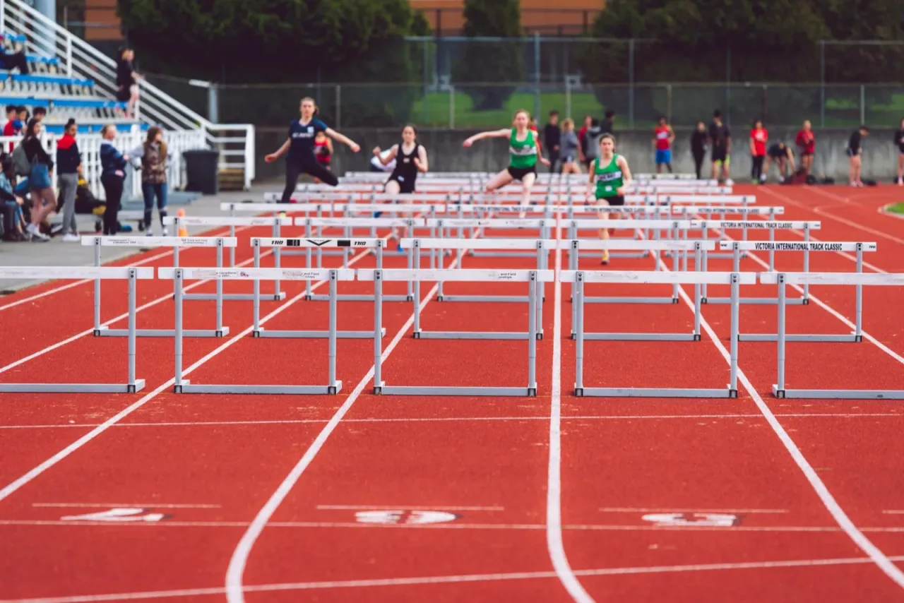 Track athletes racing hurdles on a red outdoor track surface during competition
